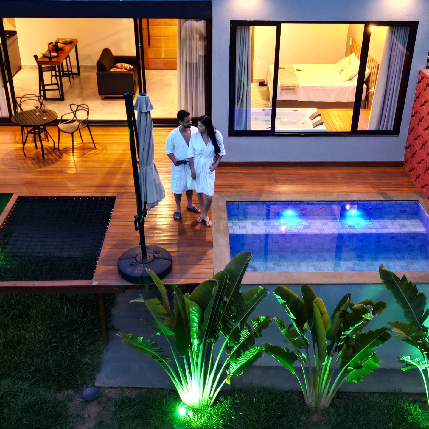 Casal de roupão ao lado da piscina iluminada à noite, com vista para o loft