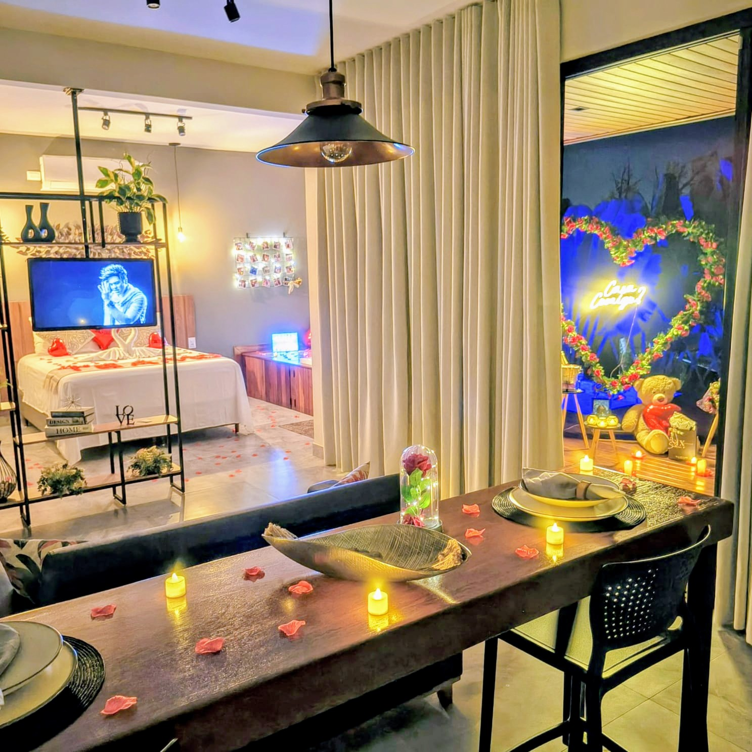 Mesa com velas e pétalas diante do coração floral iluminado com o letreiro Casa Comigo